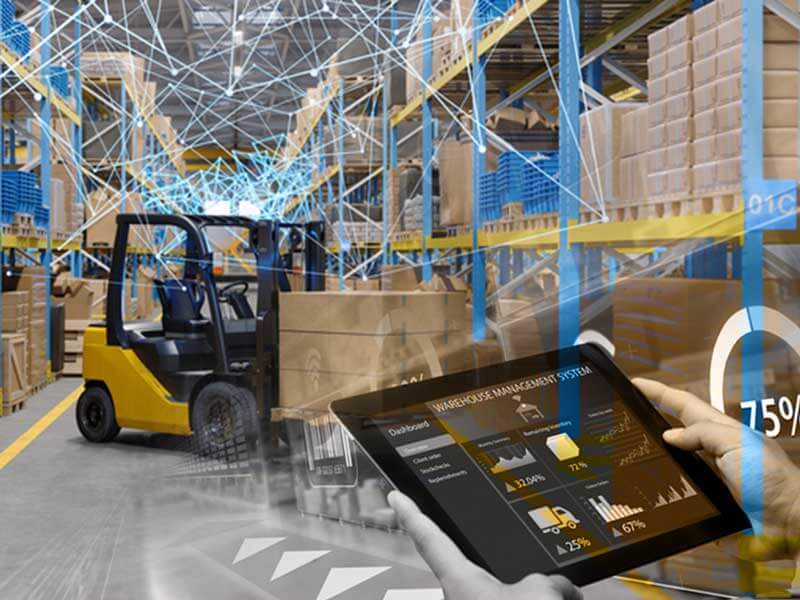 An interior view of a warehouse, with a worker in the foreground using a tablet to access the warehouse's management system. A forklift driver can be seen in the background moving a pallet off a rack.
